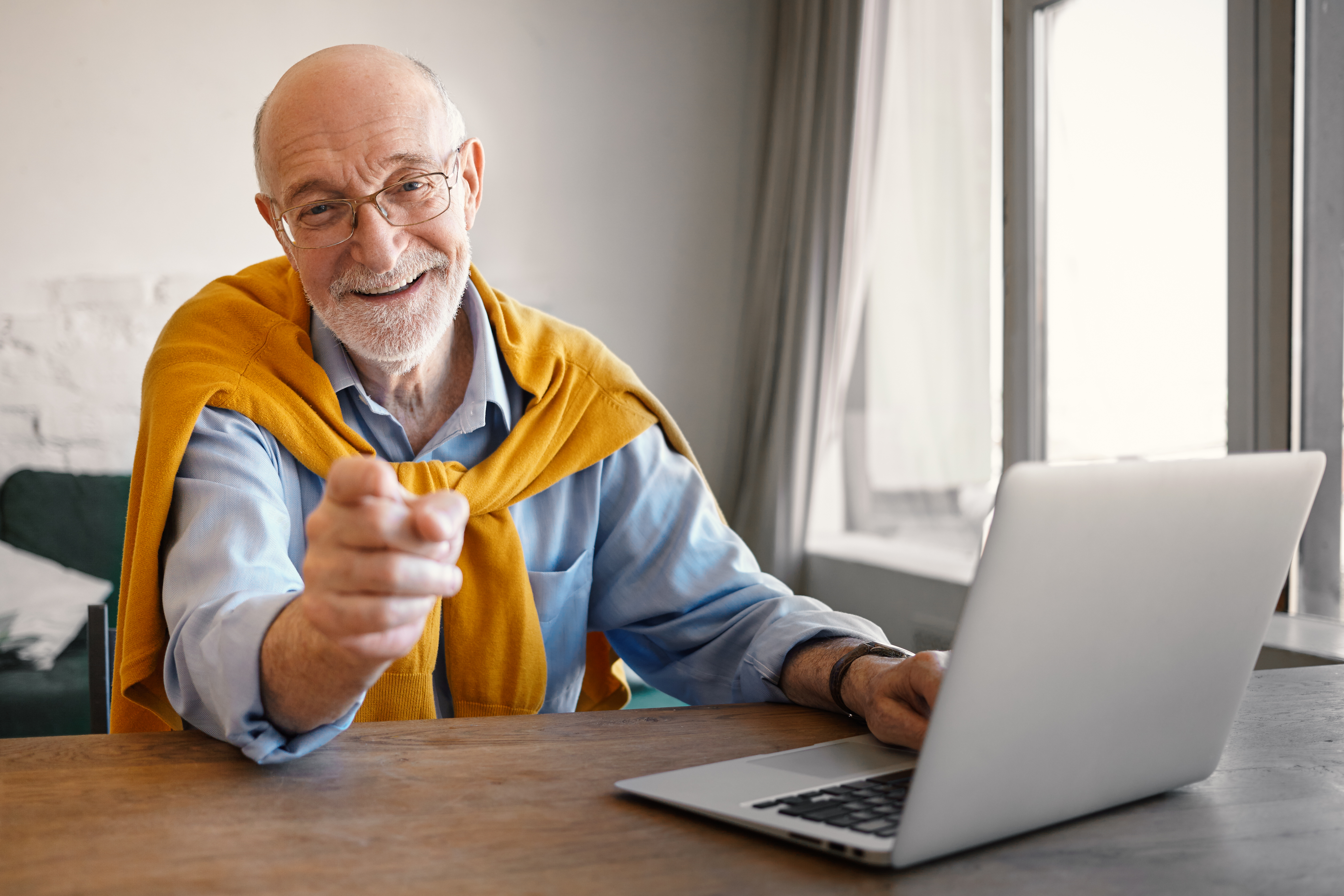 fashionable-friendly-elderly-male-recruiter-wearing-rectangular-glasses-and-elegant-clothes-working-on-portable-computer-smiling-broadly-and-pointing-finger-choosing-you-for-job-position